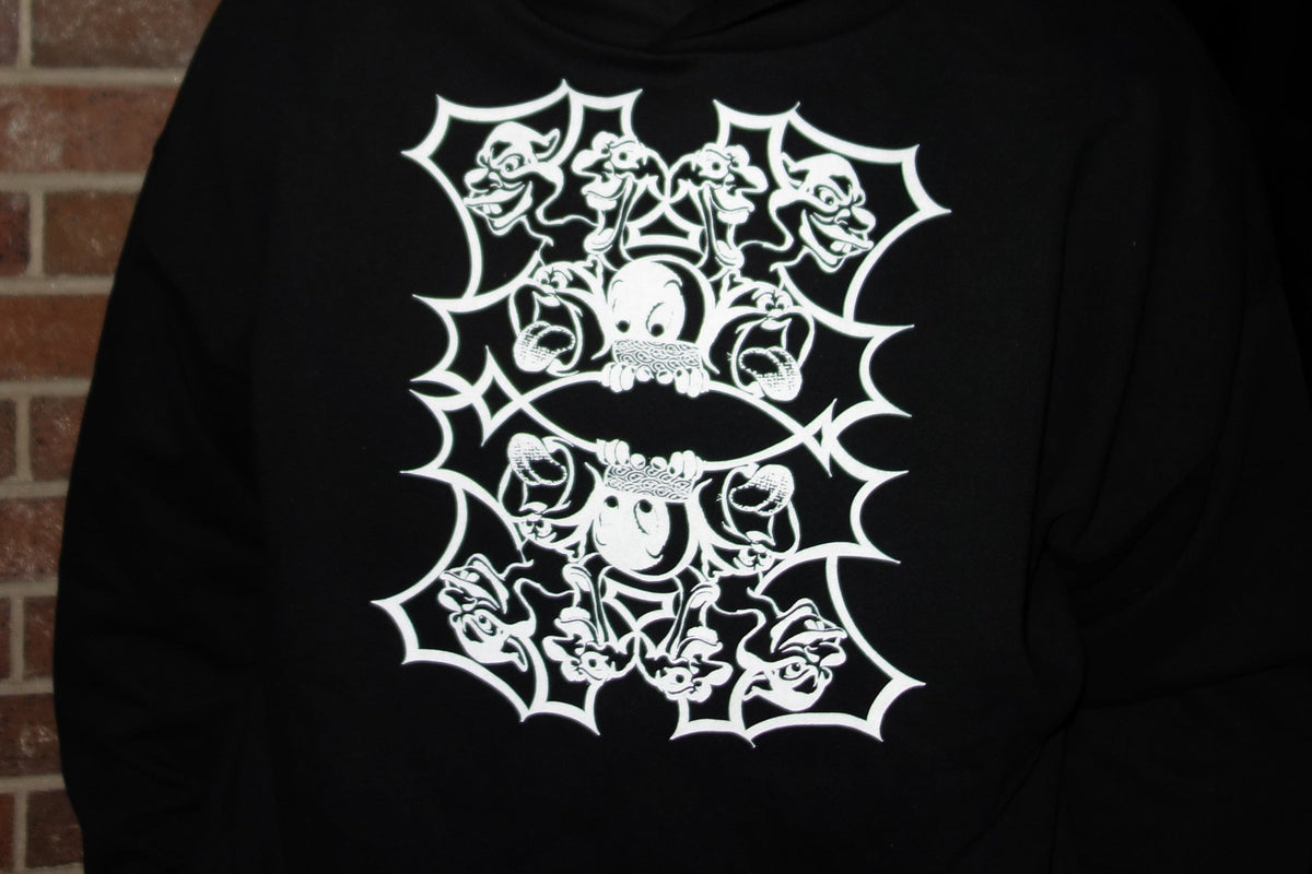 A person wearing a black pullover hoodie with a large white gothic graphic featuring skulls, bats, and roses, standing in front of a brick wall.